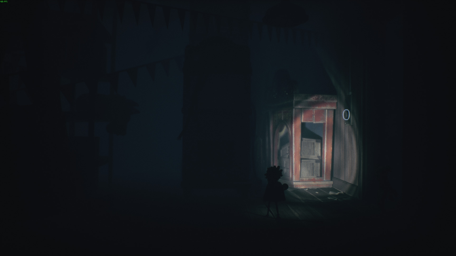 A small character approaches a red wooden booth in Little Nightmares 3, illuminated by a faint beam of light in the darkness