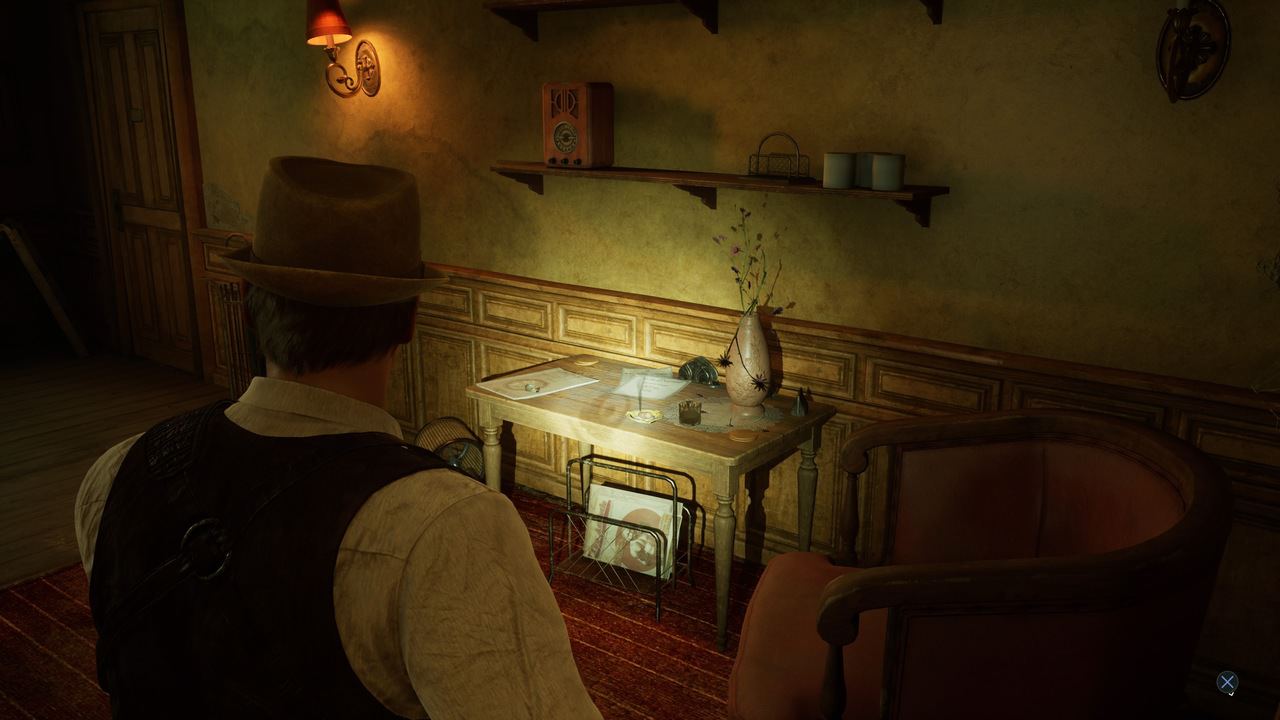 A man in a hat stands alone in the dark, in a dimly lit vintage room, looking at a table with various items including a photograph and a vase with flowers.