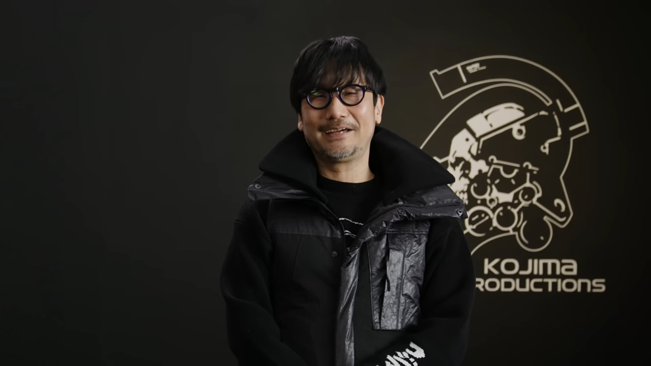 Hideo Kojima smiles against a dark backdrop with the Kojima Productions logo