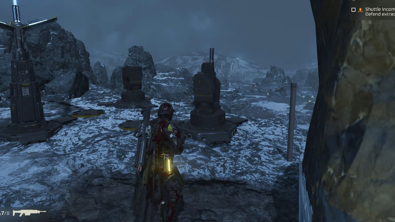 A character in a red and black suit stands in a snowy landscape near futuristic structures and rocks, under a dark, cloudy sky, as Helldivers 2 players watch expectantly.