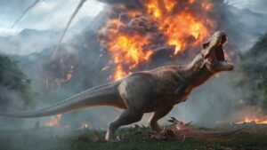 A roaring T-Rex stands on a grassy field as a volcano erupts behind it in Jurassic World Evolution 3, with fire and smoke filling the jungle sky