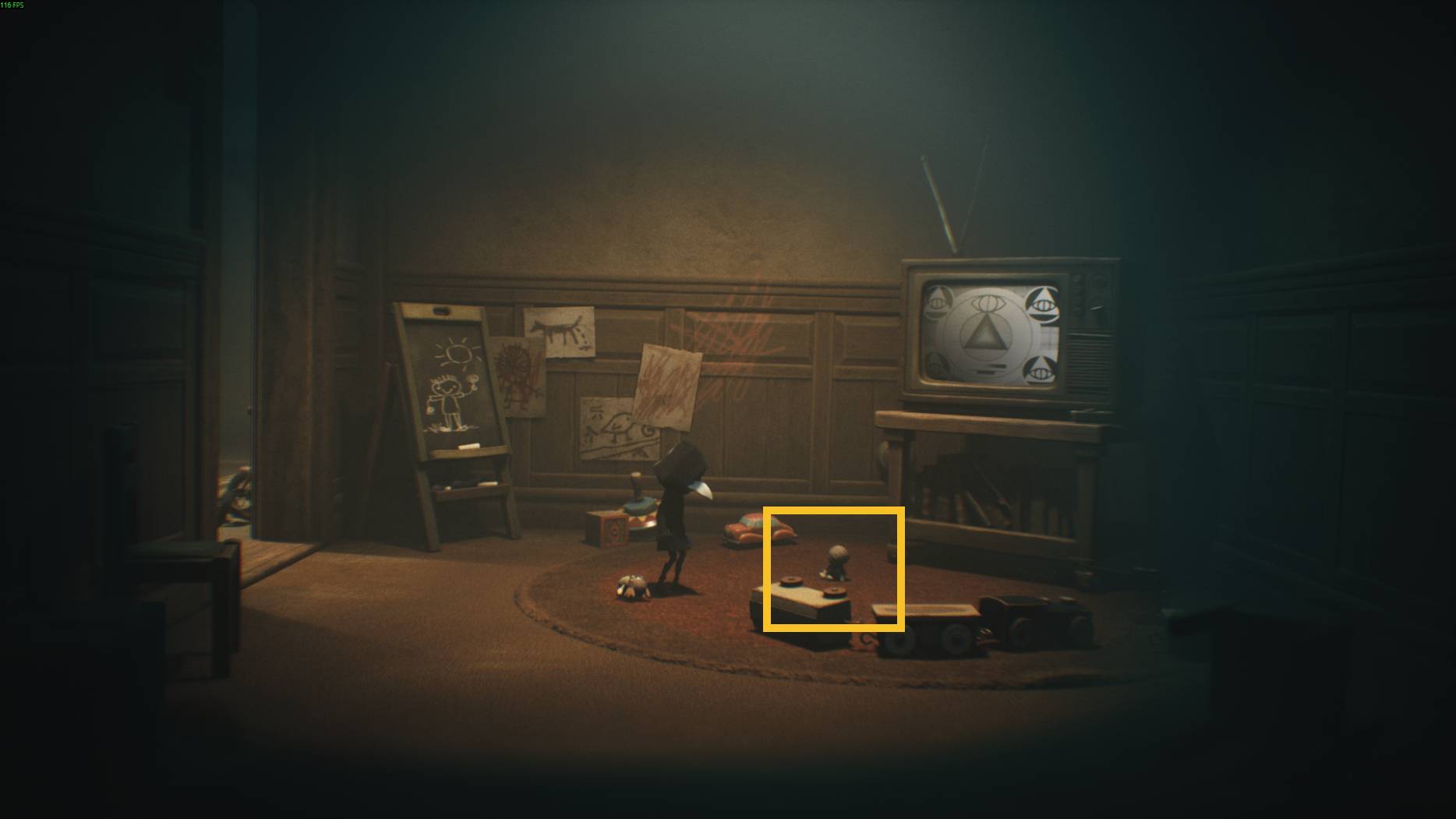 A small hidden figure stands on a rug surrounded by toys in a classroom with drawings on the wall and a TV showing strange symbols in Little Nightmares 3.