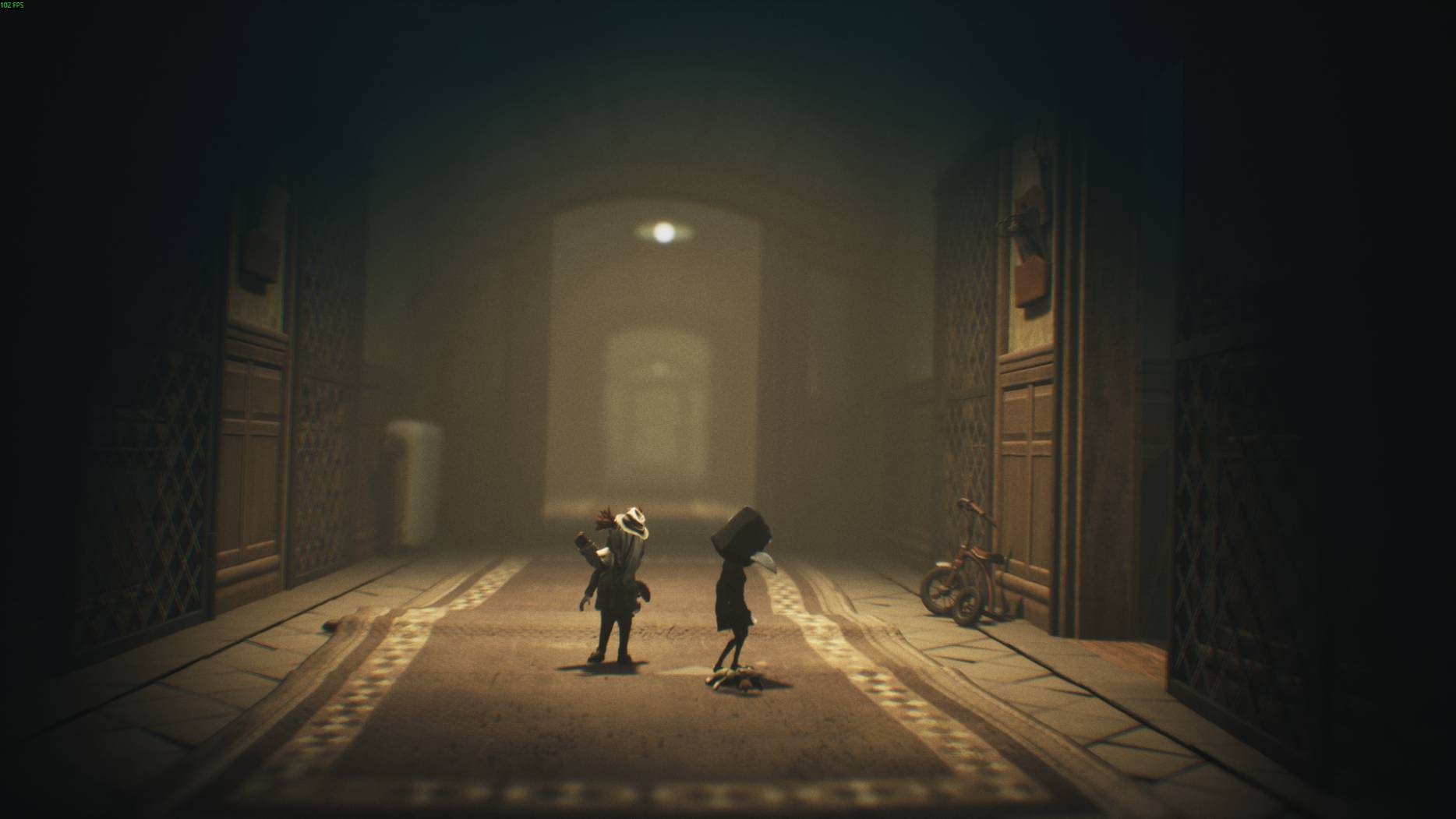 Two characters explore a long dim hallway with wooden walls and a tricycle leaning against the wall in Little Nightmares 3