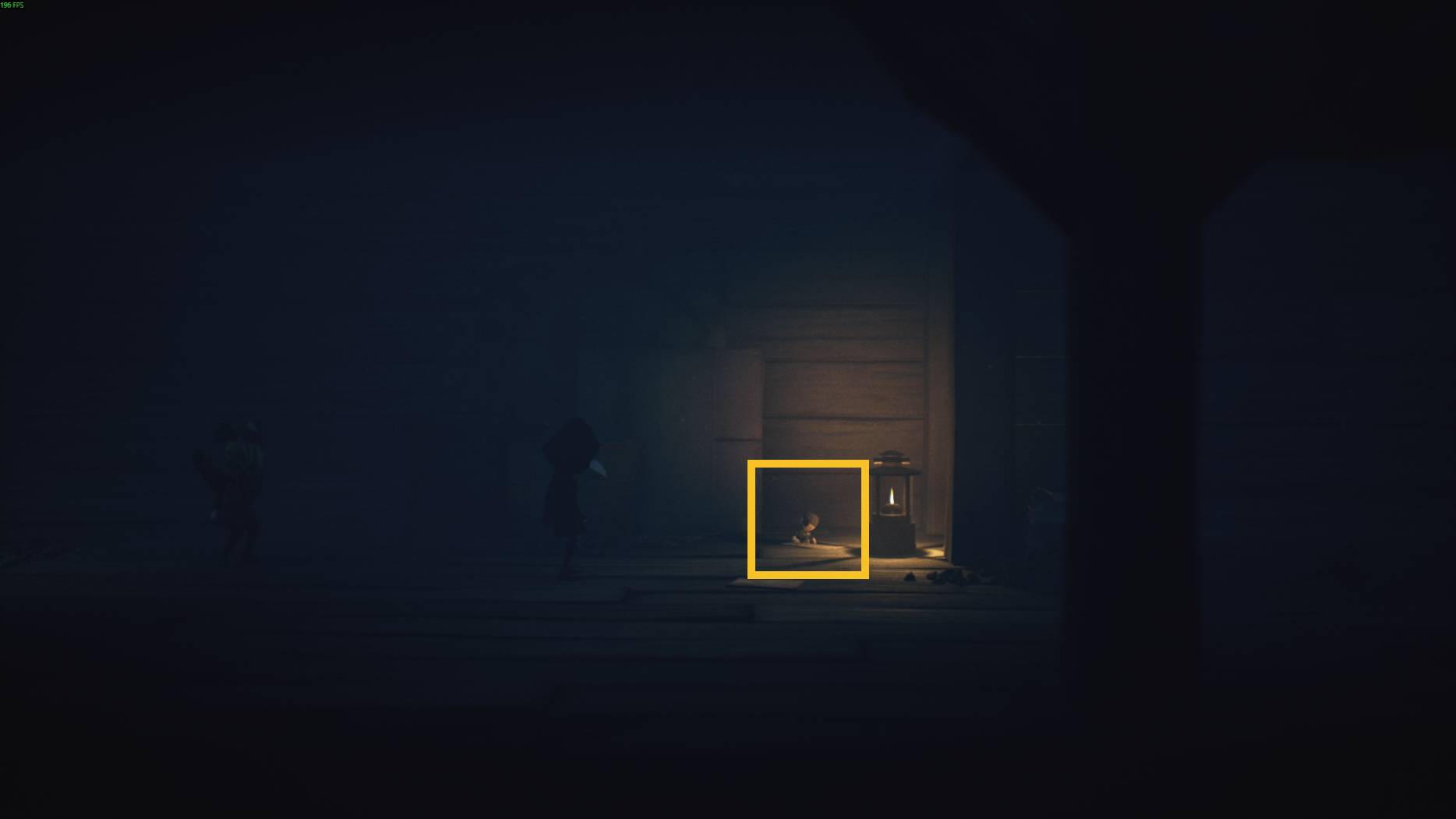 A small doll-like figure sits by a warm lantern in a dark wooden room as a hooded small character approaches in Little Nightmares 3
