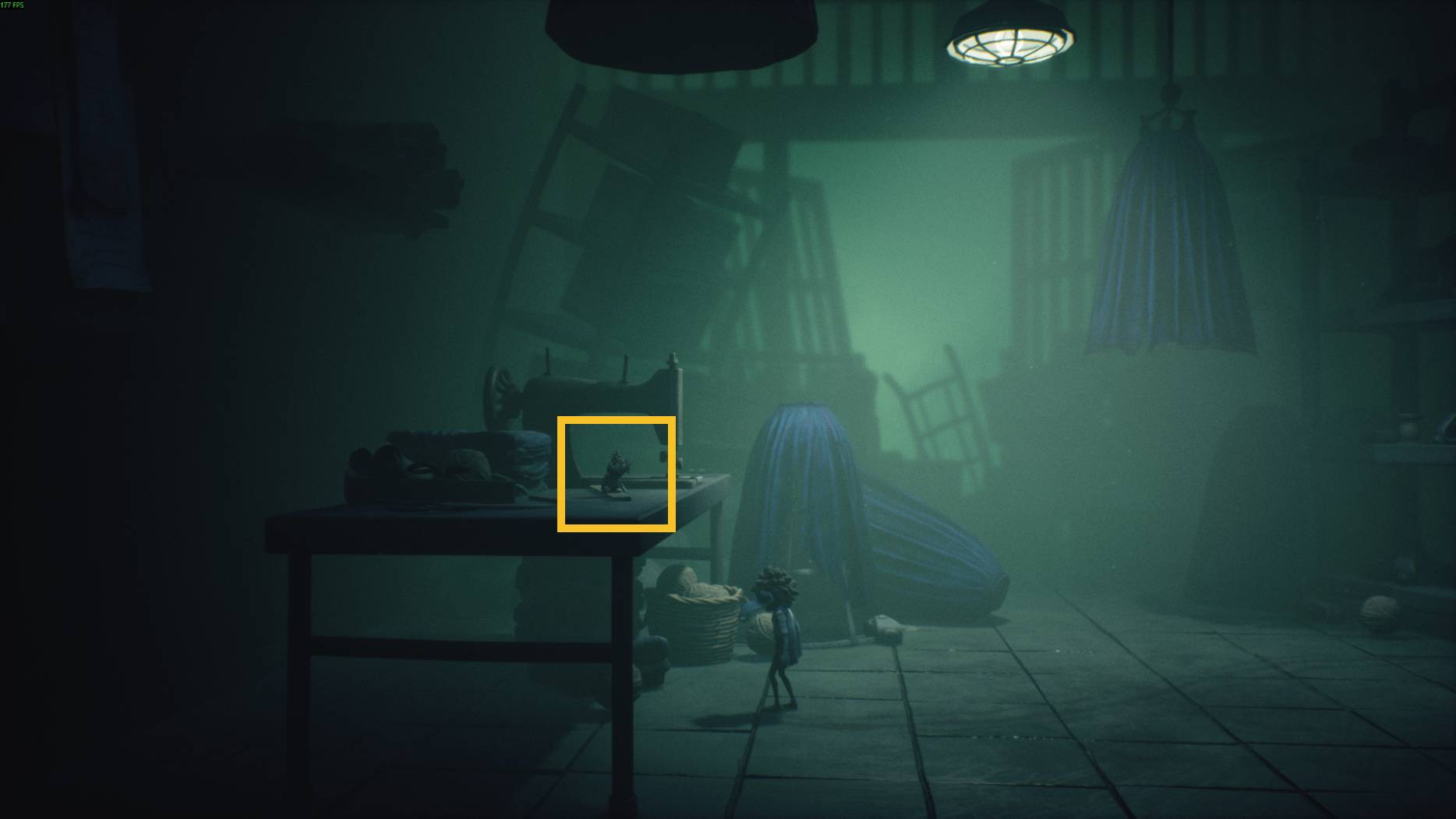 A small character stands on a sewing table in a dim workshop filled with fabric and a sewing machine in Little Nightmares 3.