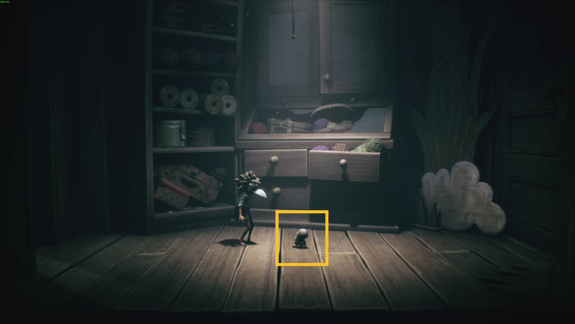 A small doll-like figure sits on the wooden floor of a sewing room filled with yarn and cabinets in Little Nightmares 3.