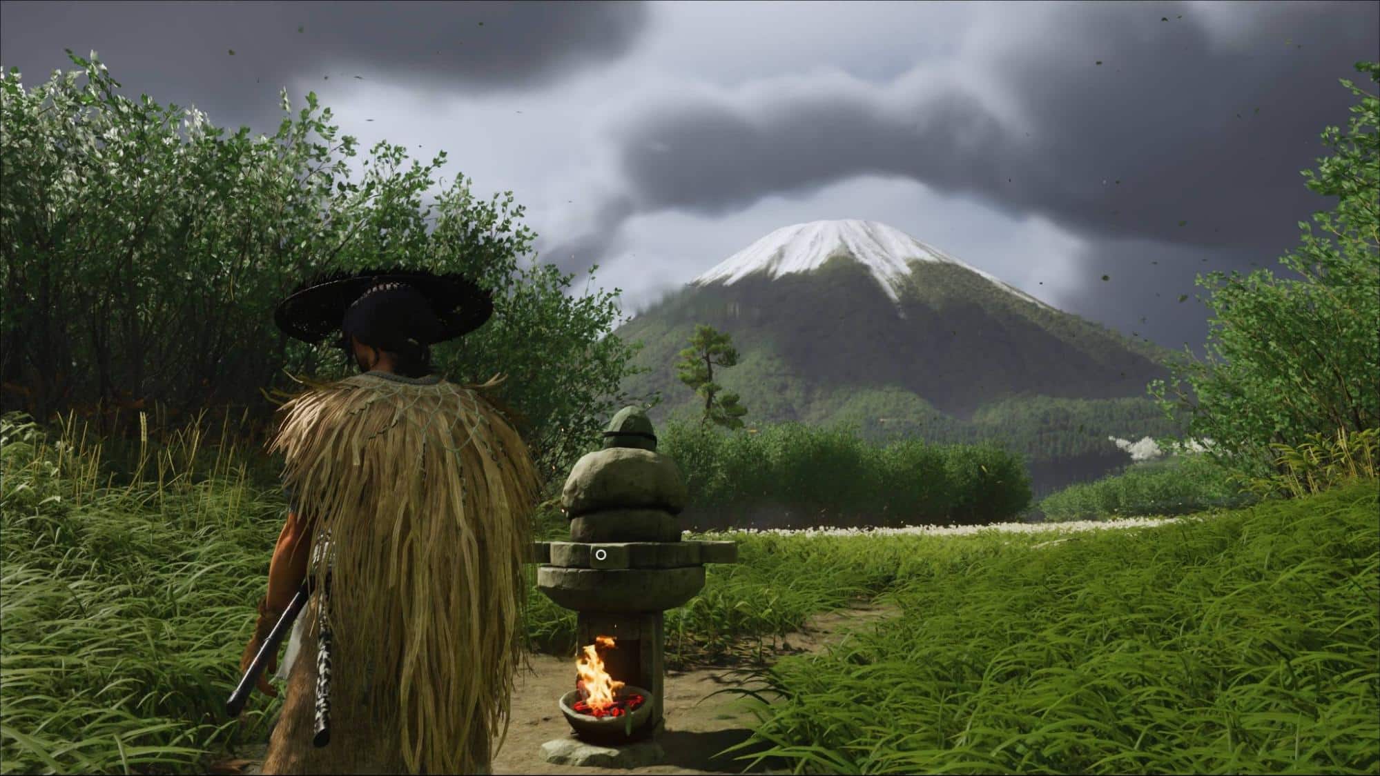 A figure in a straw cloak and wide-brimmed hat stands beside a stone lantern with a small fire, facing a snow-capped mountain under stormy skies in Ghost of Yotei