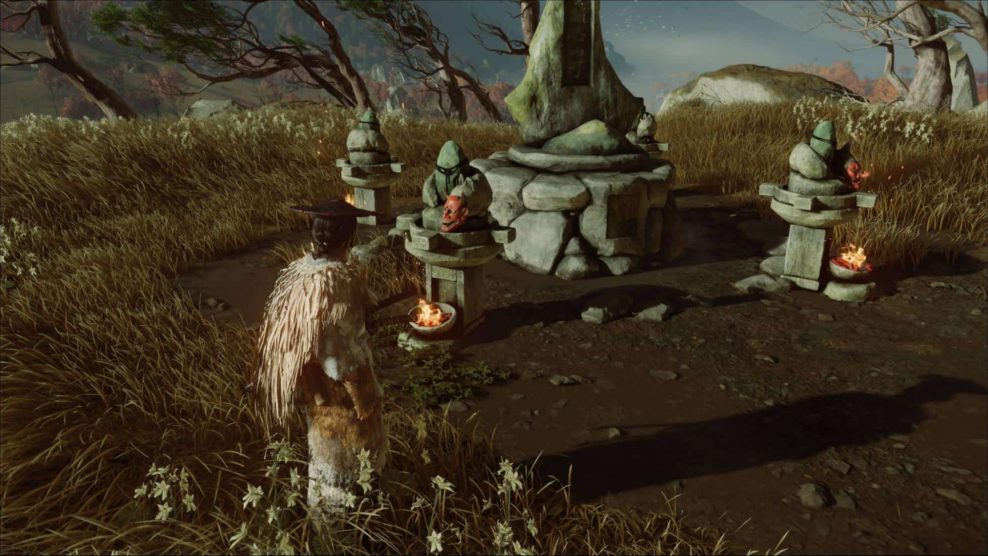 A figure in a straw cloak stands before a stone shrine with small statues, surrounded by tall grass and twisted trees under an overcast sky in Ghost of Yotei