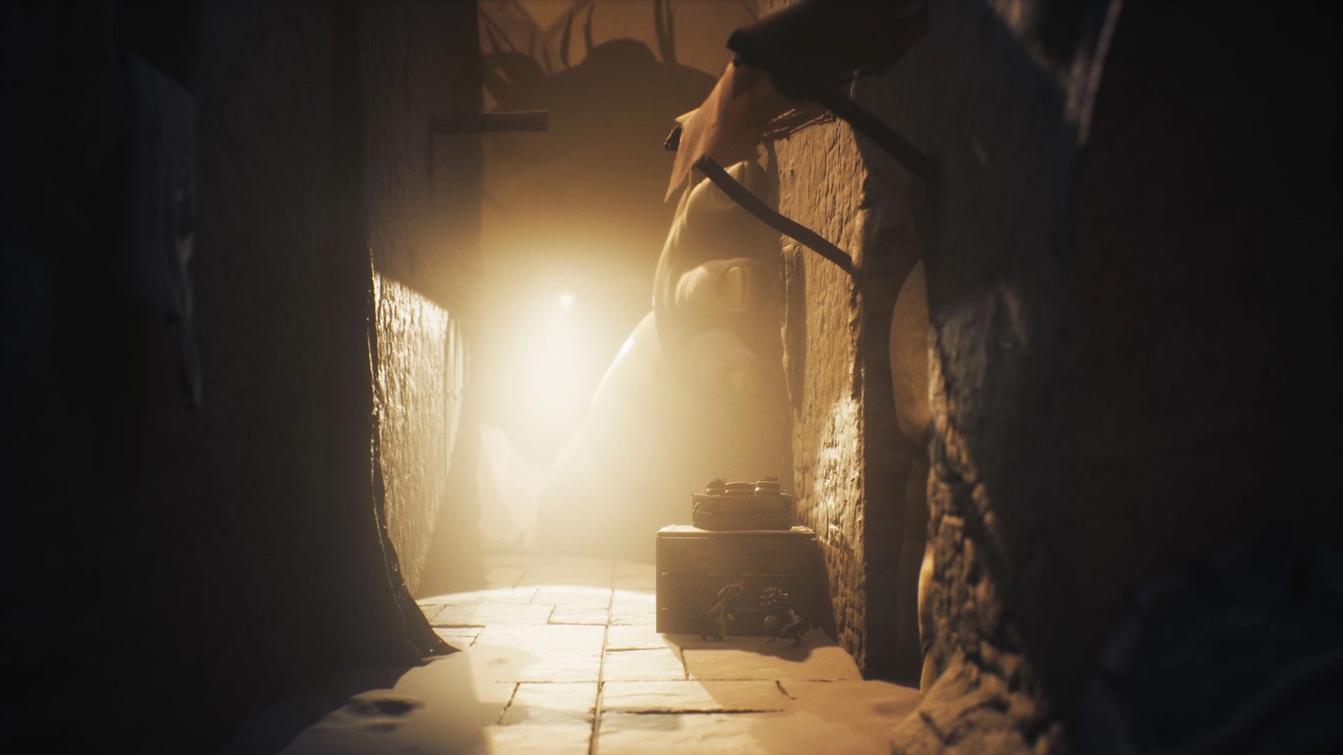 Low and Alone hide in a dim hallway from a giant creature’s hand in Little Nightmares 3