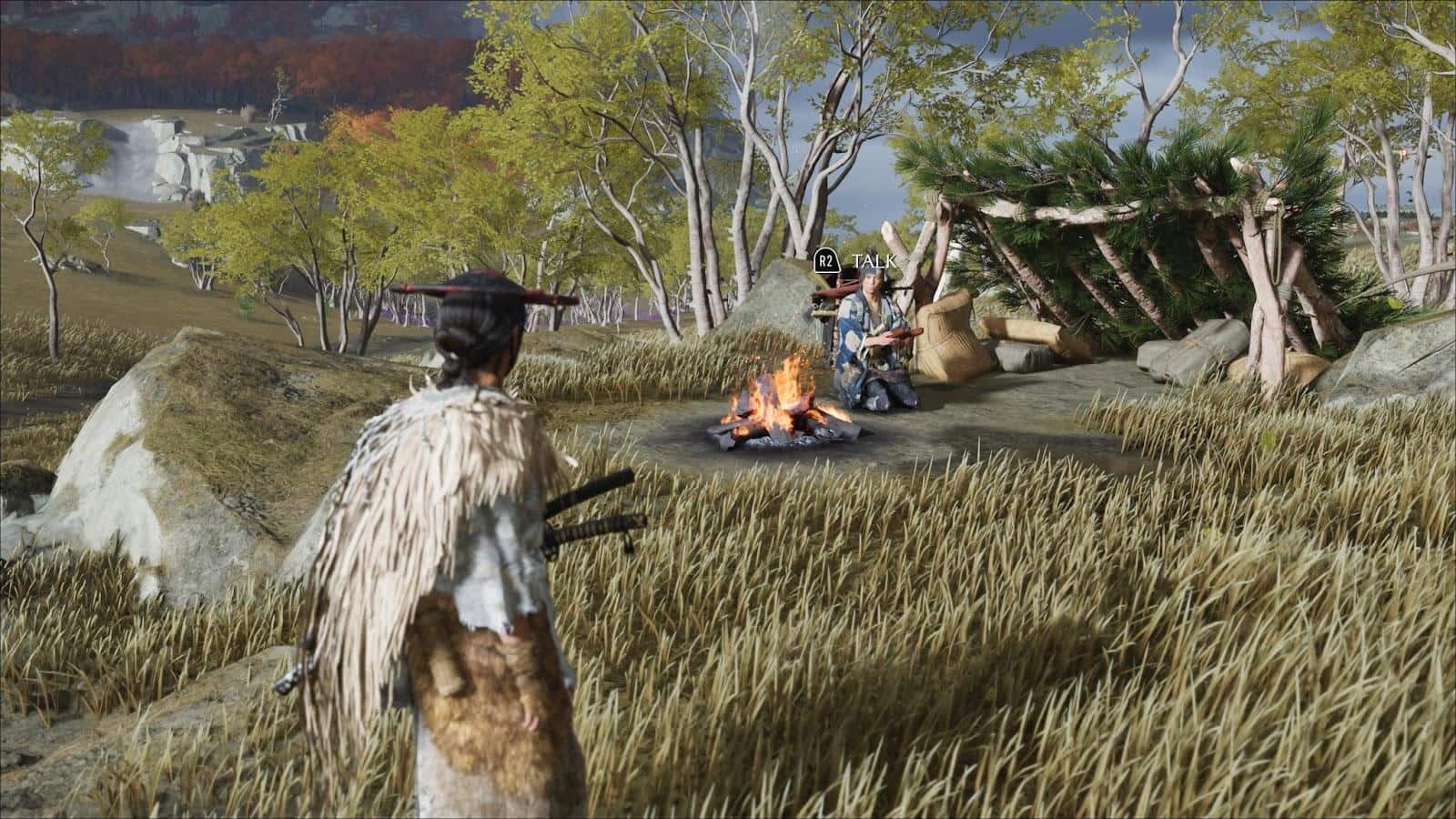 Ghost of Yotei game scene. A person in a fur cloak and conical hat stands in a grassy field, approaching another person seated by a campfire near a shelter of branches.