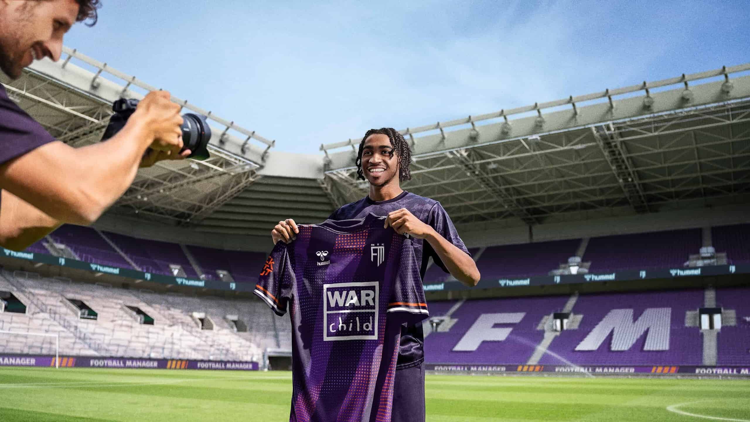Football Manager 2024: A footballer holding up a jersey in an empty stadium while a man takes his picture.