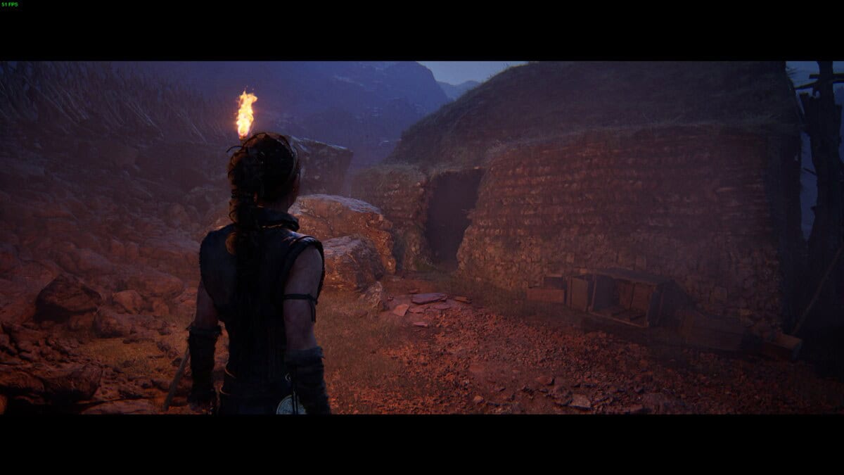 Hellblade 2 Hidden Faces locations: Senua stands with a torch in hand, facing a dark, stone-built house.