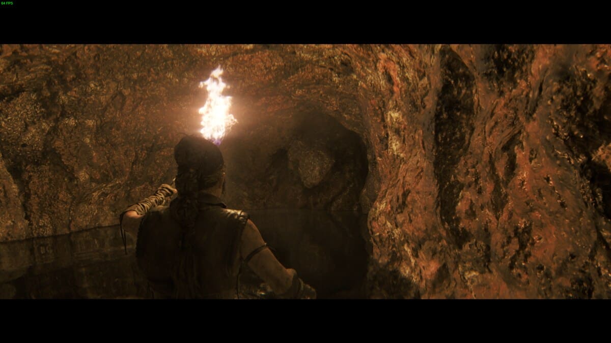 Hellblade 2 Hidden Faces locations: Senua holding a burning torch, standing in a rocky, dimly lit cave.