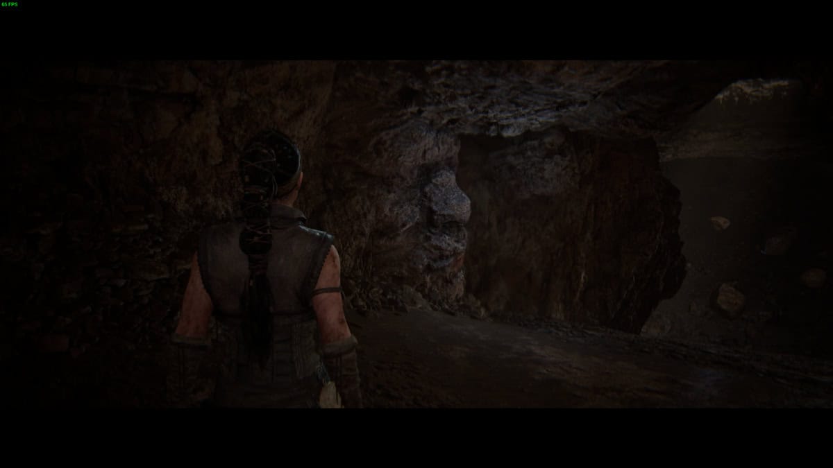 Hellblade 2 Hidden Faces locations: Senua stands in a dark, rocky cave, looking toward an exit illuminated by dim light.