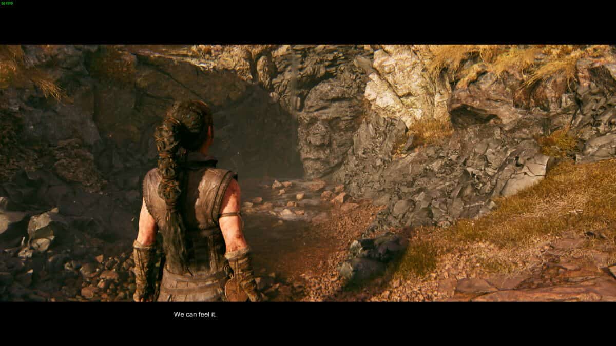 Hellblade 2 Hidden Faces: Senua stands before a rocky cave entrance with a face etched in the rock.