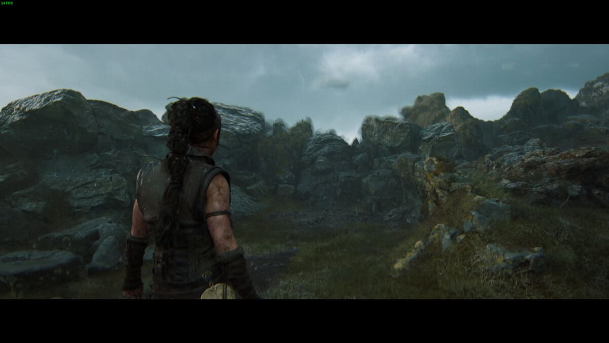 Hellblade 2 Hidden Faces: Senua standing in a rocky landscape under a cloudy sky with a face carved into the rocks ahead.