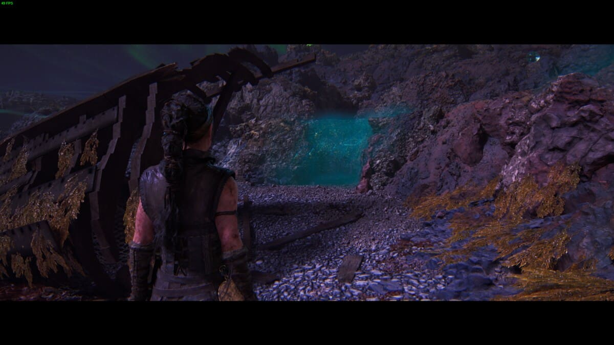 Hellblade 2 Hidden Faces: Senua stands in front of a partially submerged, decayed shipwreck, surrounded by rocky terrain and illuminated with a blue-green light.