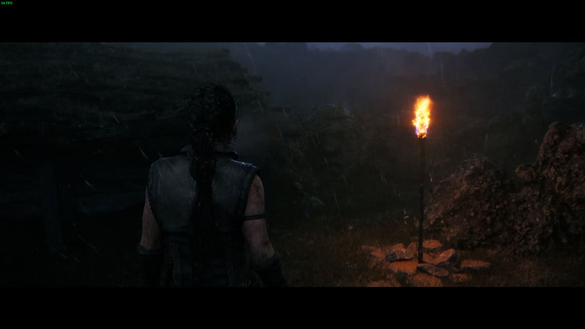 Hellblade 2 Hidden Faces: Senua stands in the rain facing a lit torch on a stand, with a gloomy village with a face carved into the rocks ahead.