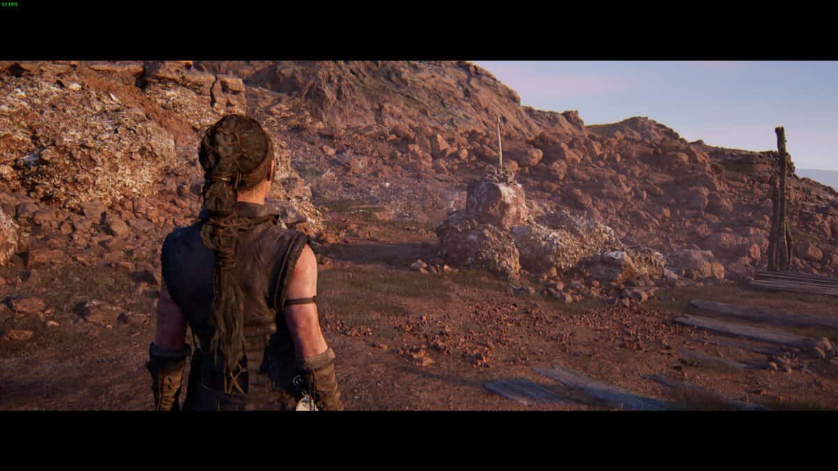 Hellblade 2 Hidden Faces locations: Senua stands on a rocky terrain, looking towards a rocky hill under a clear sky.