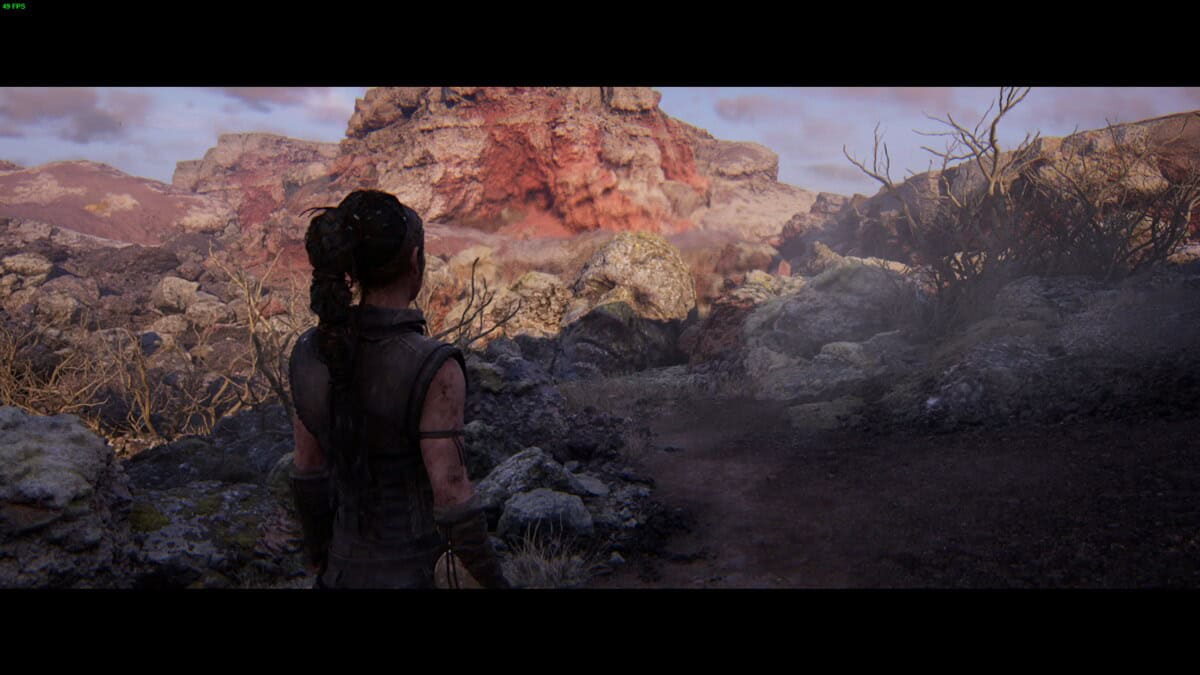 Hellblade 2 Hidden Faces locations: Senua stands on a rocky path, facing a rugged landscape with a large, red rock formation in the distance under a cloudy sky.