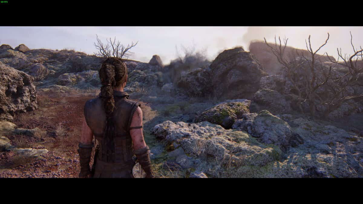 Hellblade 2 Hidden Faces locations: Senua stands on a rocky landscape with sparse vegetation, looking towards a face etched into the rocks ahead.