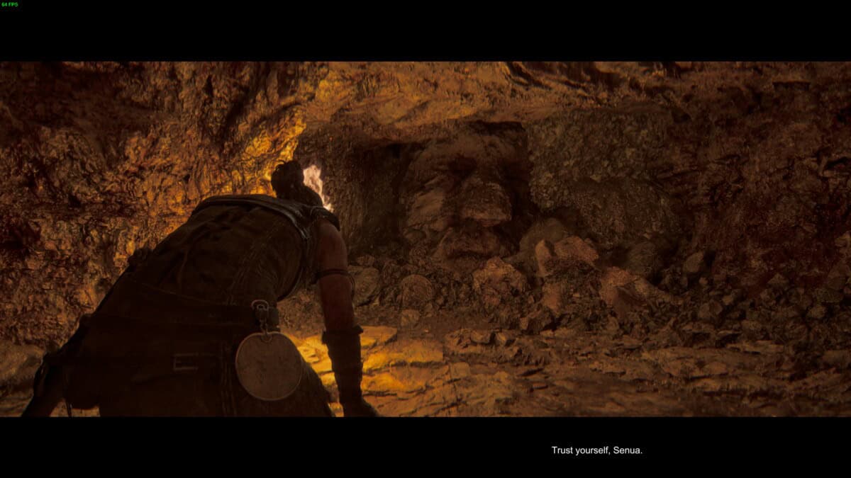 Hellblade 2 Hidden Faces locations: Senua holding a torch kneels in a dimly lit, rocky cave with a faintly human-like face formed in the rock.