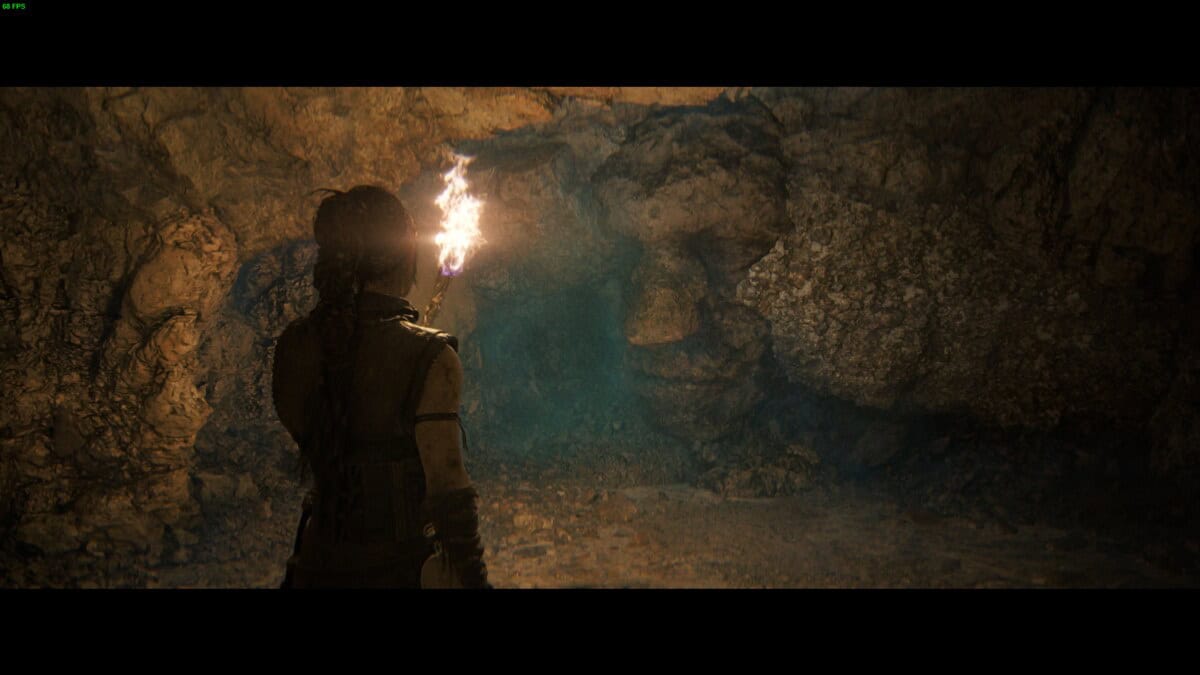 Hellblade 2 Hidden Faces locations: Senua holding a lit torch in an underground cave with rough, rocky walls shaped like a face.