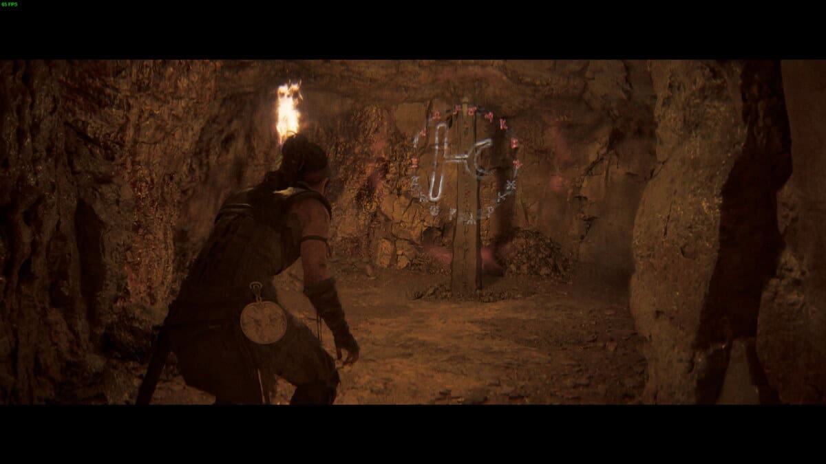 Hellblade 2 Lorestangir locations: Senua holding a torch is in a cave, facing a circular, glowing rune.