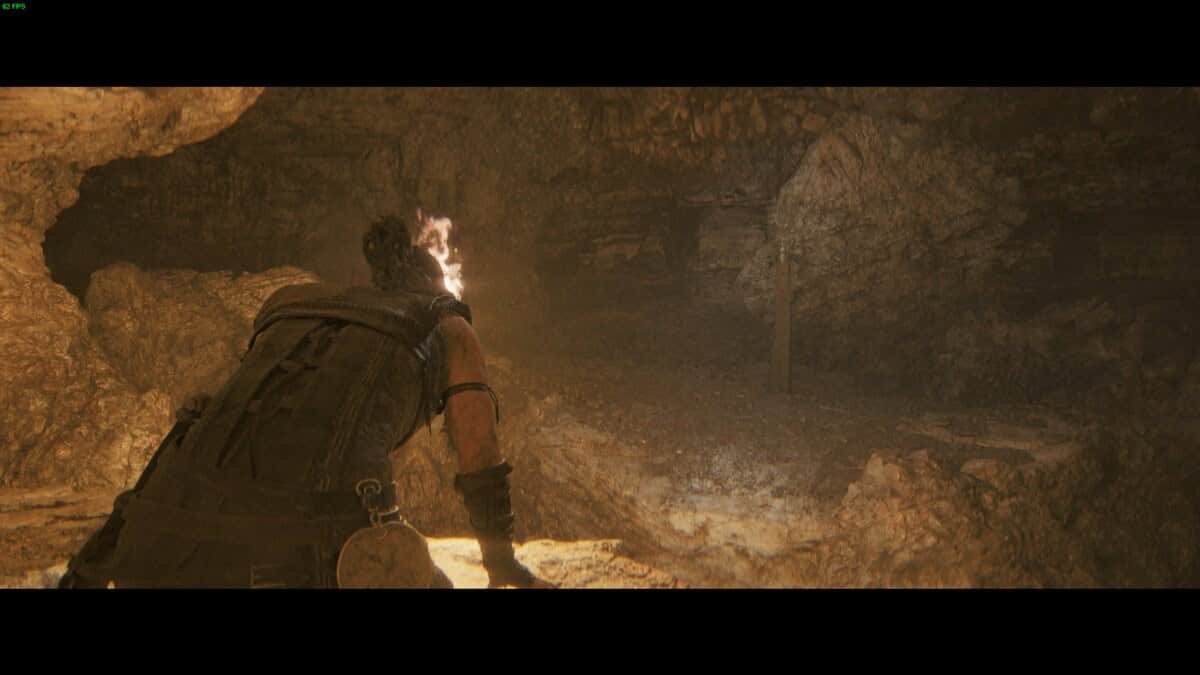 Hellblade 2 Lorestangir locations: Senua crawling through a dimly lit, rocky cave with a torch in hand.