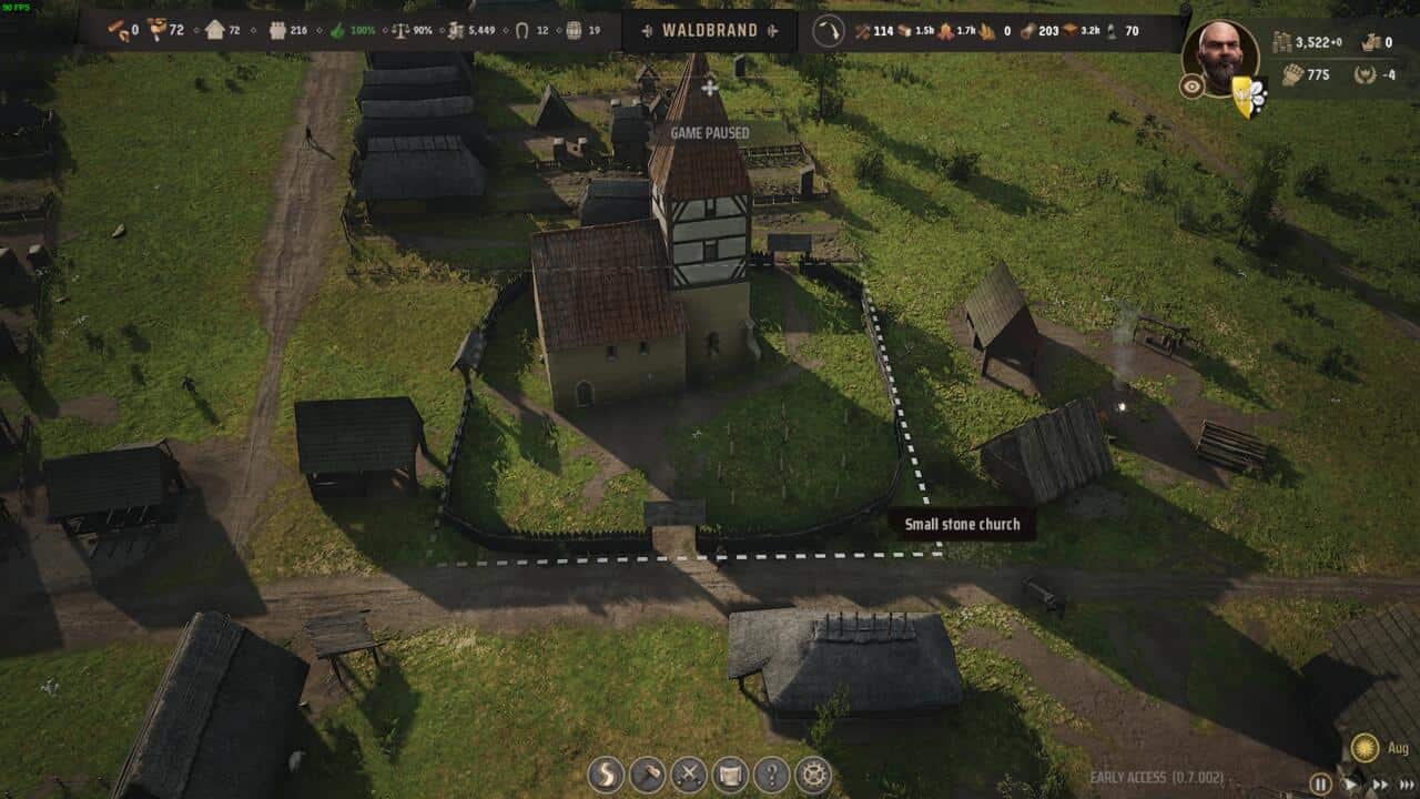 Manor Lords buildings: Overhead view of a medieval village featuring Manor Lords buildings, including a small stone church and thatched-roof houses, surrounded by palisade fencing, with game interface elements visible.