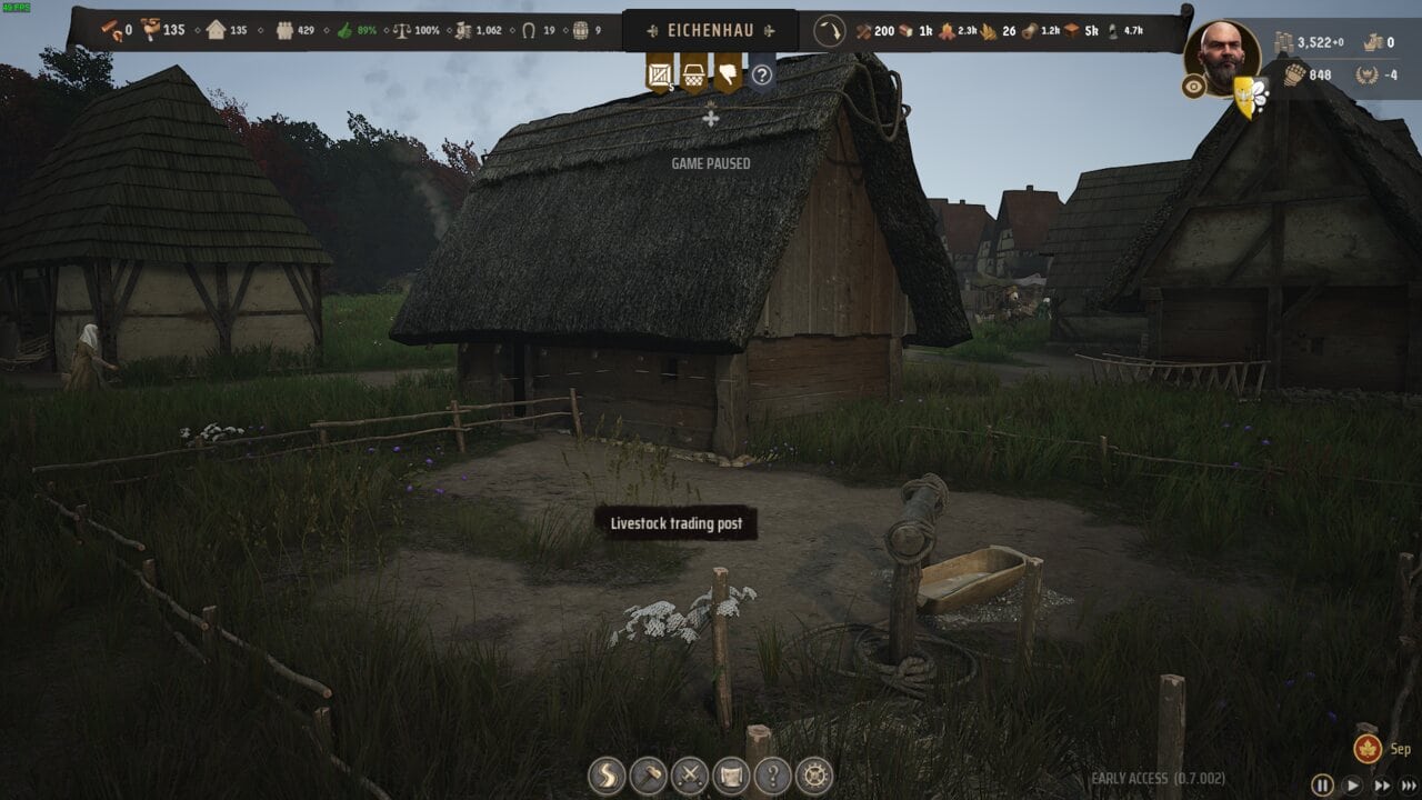 Manor Lords buildings: Screenshot of a village scene in Manor Lords, featuring thatched-roof buildings and a livestock trading post, with game interface icons at the top.