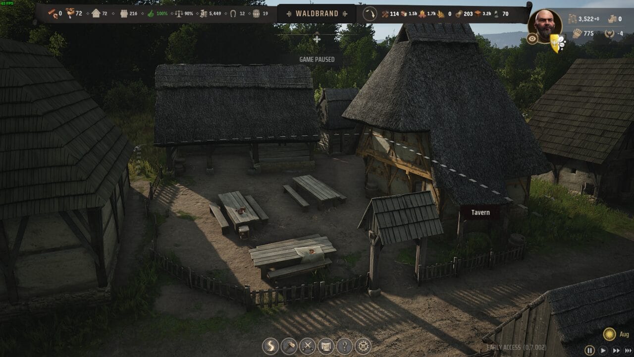 Manor Lords buildings: A paused medieval village scene from the video game Manor Lords, displaying thatched-roof huts, dirt paths, and digital interface elements.