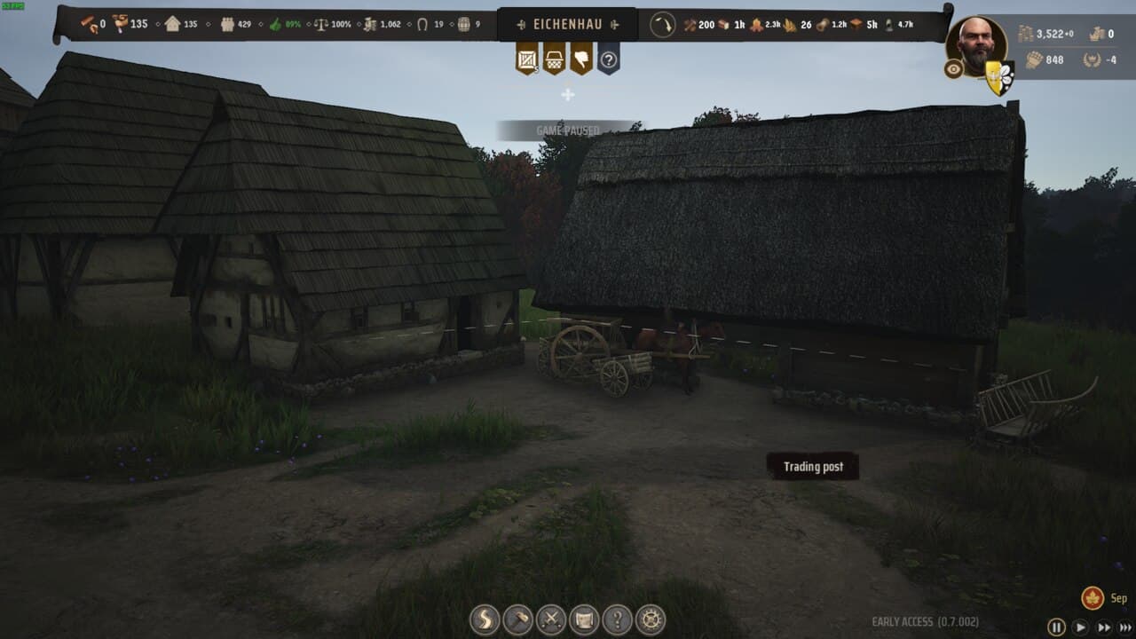 Manor Lords buildings: Screenshot from a video game showing a medieval village with two thatched-roof cottages, a horse-drawn cart, and a Manor Lords trading post sign under an overcast sky.