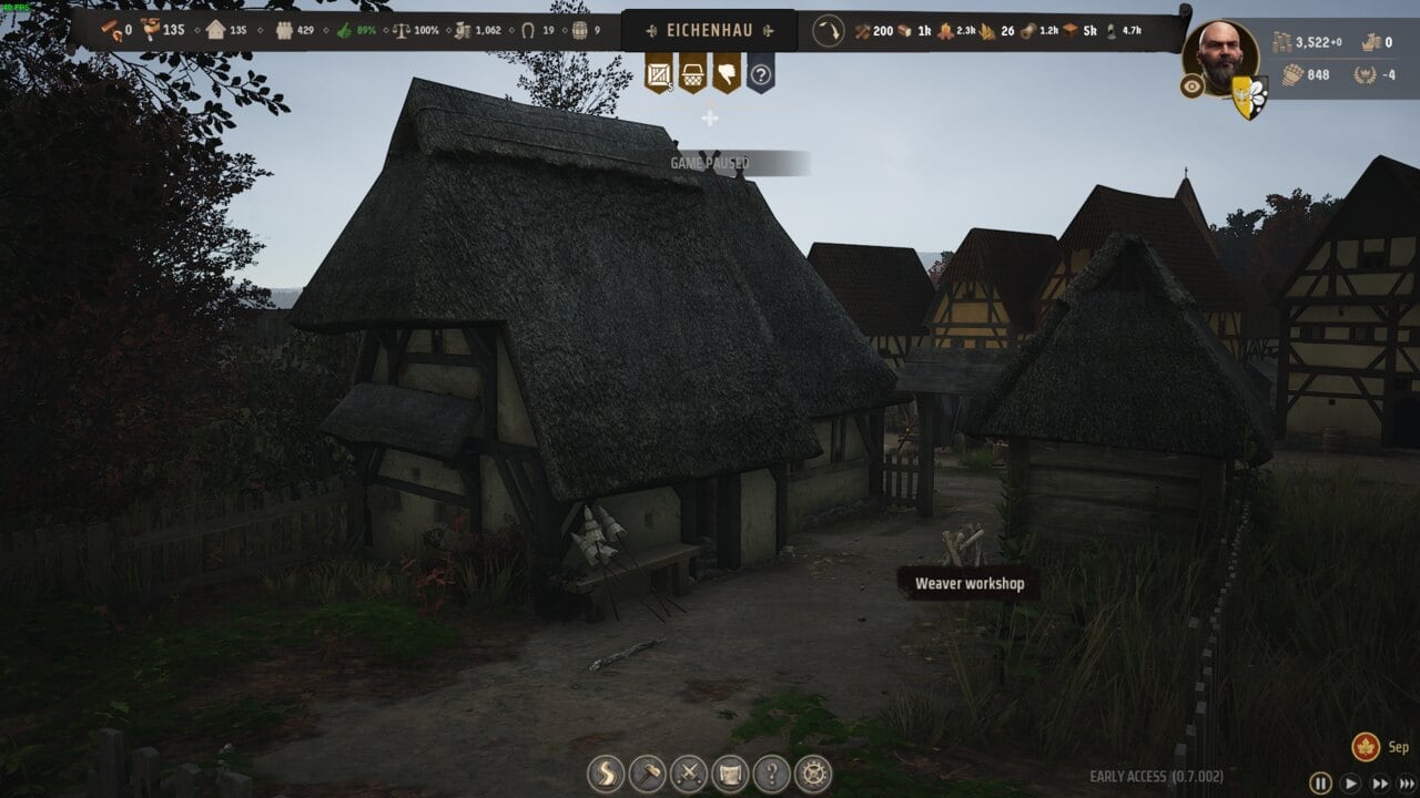 Manor Lords buildings: A screenshot from a video game showing a medieval village with detailed Manor Lords buildings including a weaver workshop under a thatched roof, indicating a focus on historical craftsmanship.