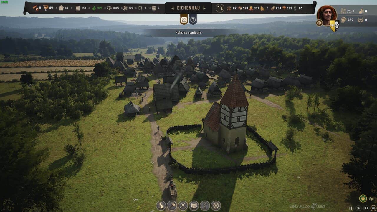Manor Lords review: aerial view of a medieval village with a large half-timbered church surrounded by smaller structures, green fields, and trees under a clear sky.