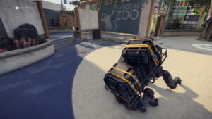 A player in XDefiant maneuvers a crouched character near a cart stacked with crates in an outdoor zoo setting, complete with signage, pathways, and trees.