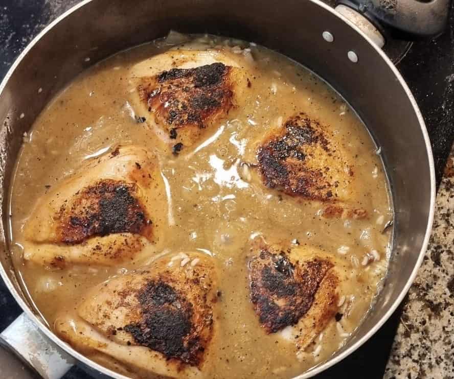 Chicken thighs in a pot submerged in chicken stock, with rice, onions, and garlic below.